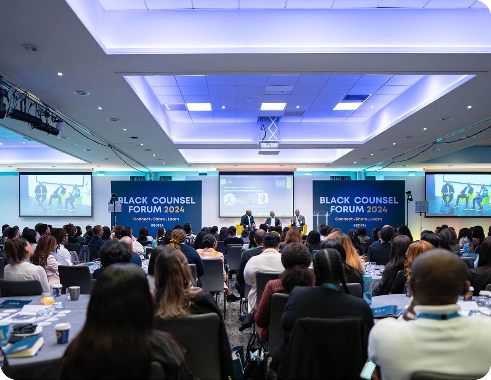 A large group of people are seated in a conference room, watching a presentation on a large screen. The audience is engaged and focused on the speaker. The room is equipped with a projector and a laptop, and there are several chairs and dining tables scattered throughout the space. The attendees are dressed in business attire, indicating a professional setting. AI generated content
