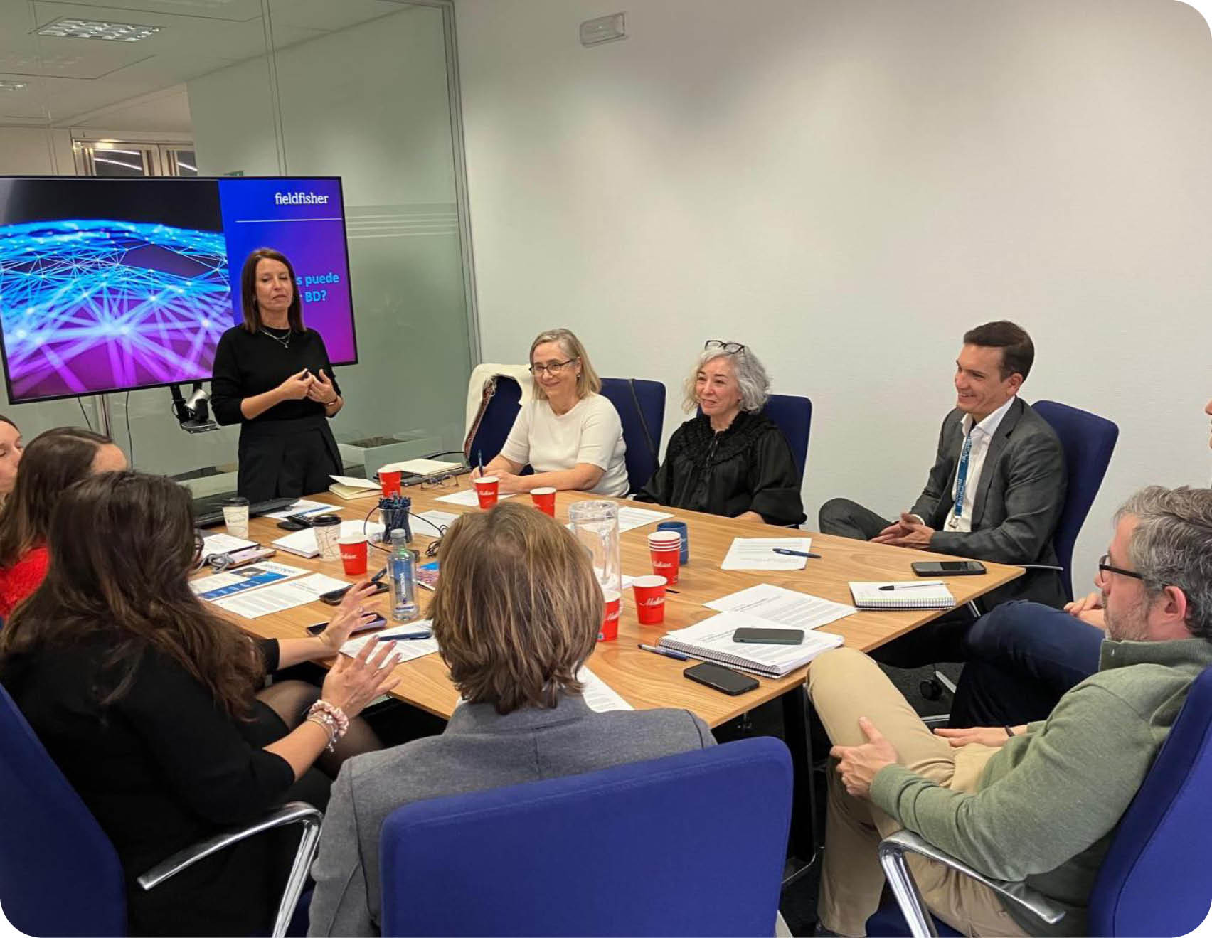 A group of people are sitting around a table, engaged in a meeting. There are several cups and a cell phone on the table. A TV is visible in the background. AI generated content