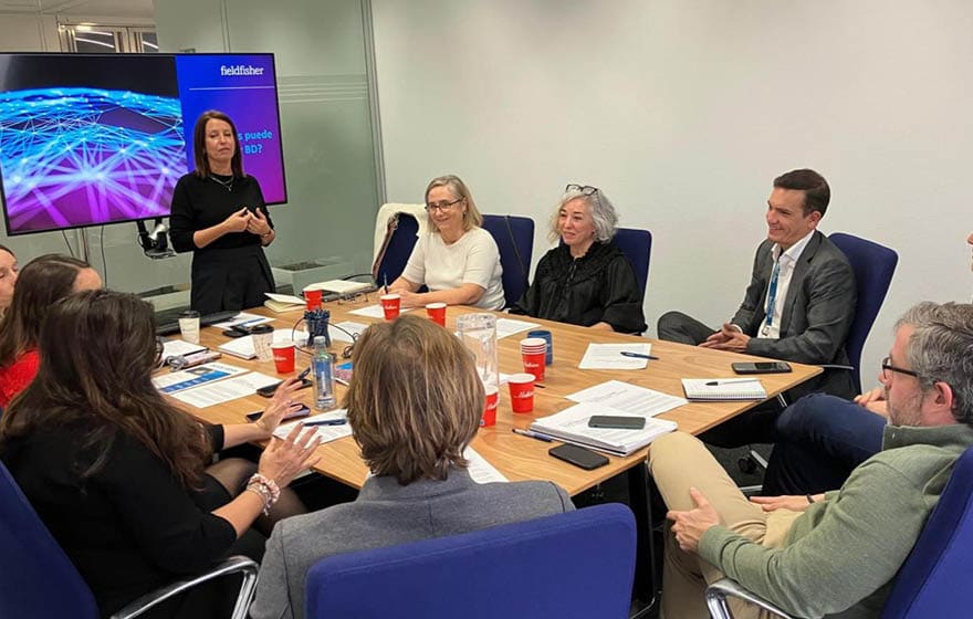 A group of people are sitting around a table, engaged in a meeting. There are several cups and a cell phone on the table. A TV is visible in the background. AI generated content
