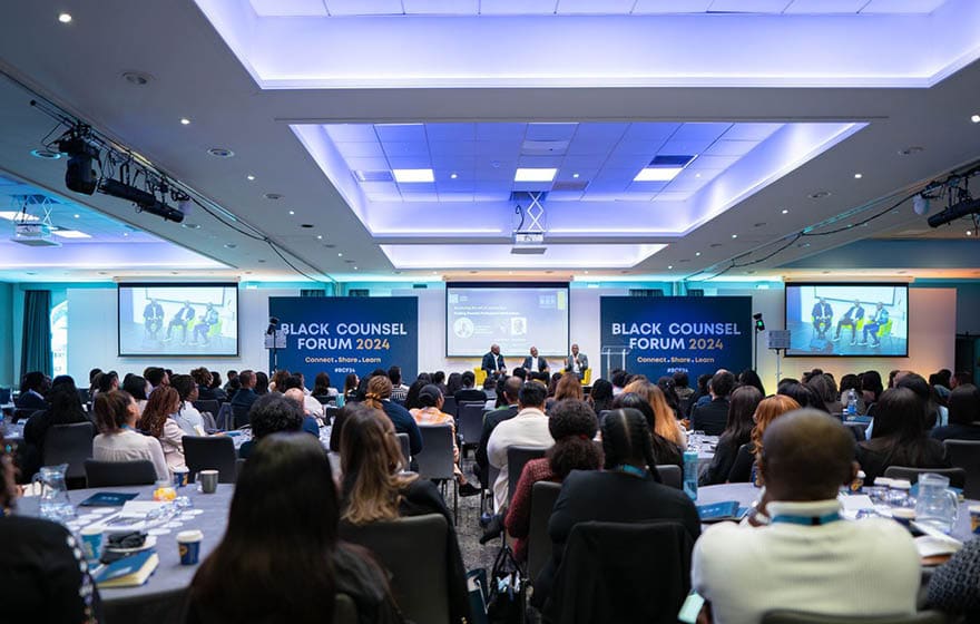 A large group of people are seated in a conference room, watching a presentation on a large screen. The audience is engaged and focused on the speaker. The room is equipped with a projector and a laptop, and there are several chairs and dining tables scattered throughout the space. The attendees are dressed in business attire, indicating a professional setting. AI generated content