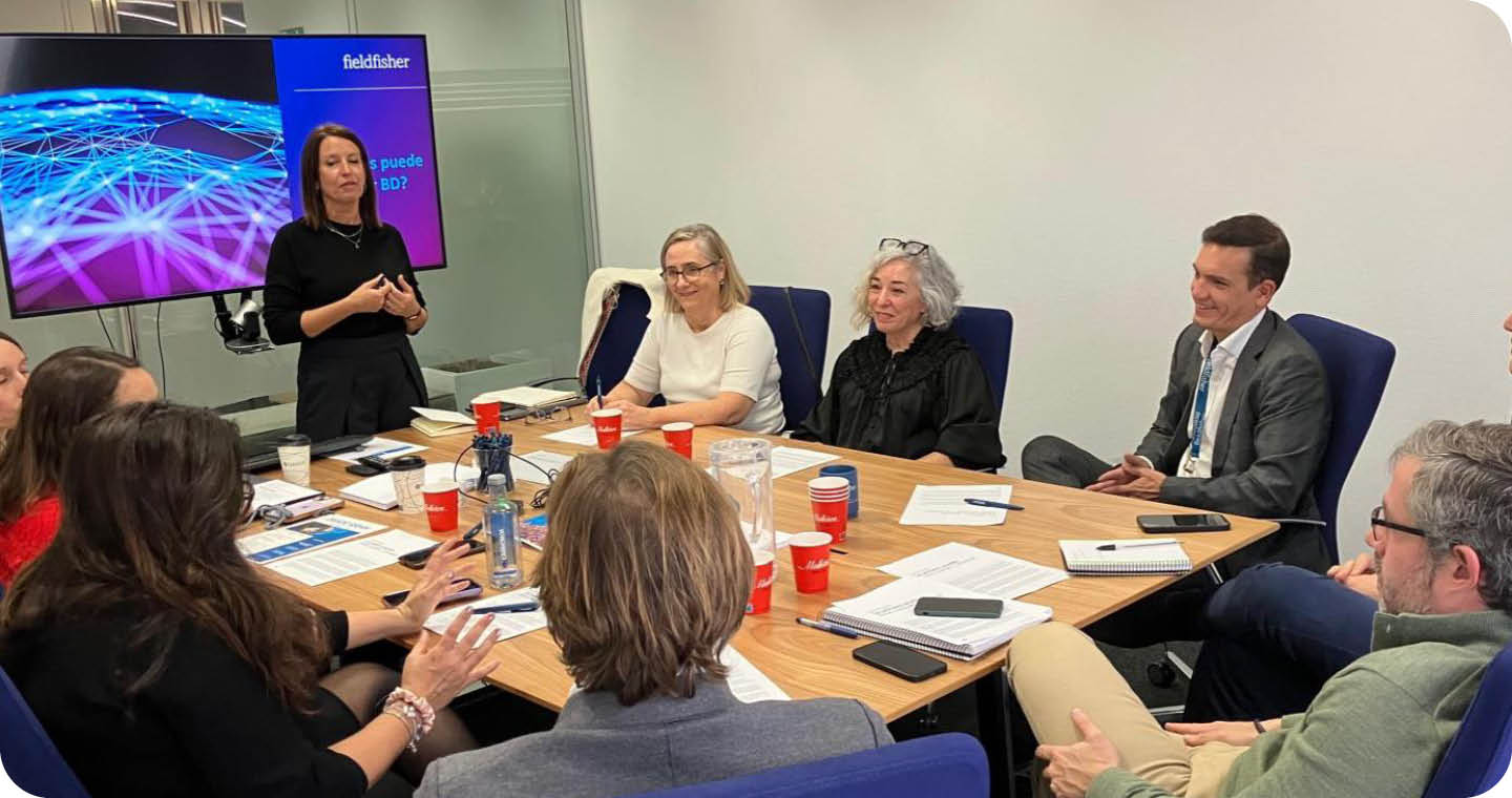 A group of people are sitting around a table, engaged in a meeting. There are several cups and a cell phone on the table. A TV is visible in the background. AI generated content